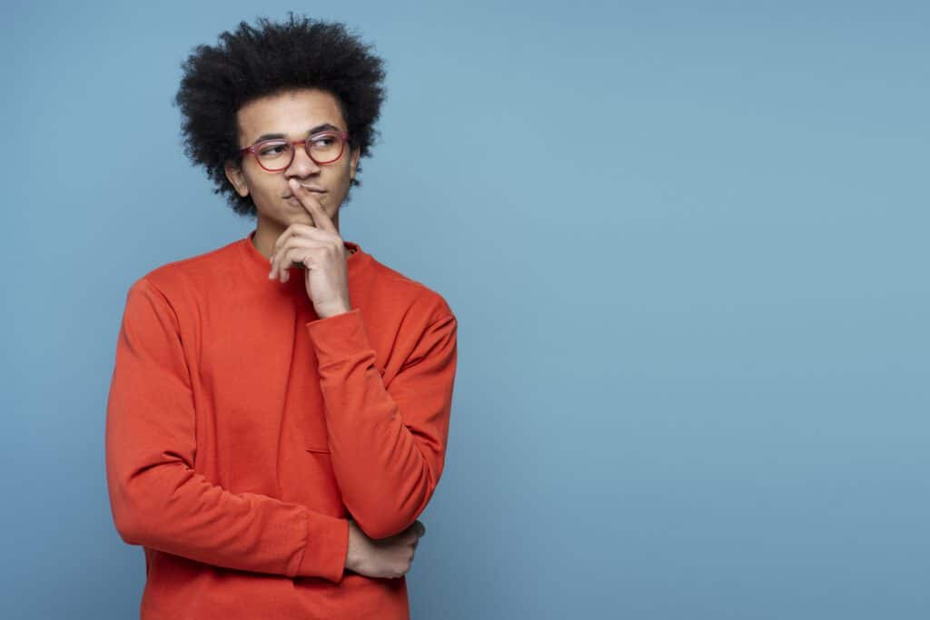 A young, pensive African American student wearing eyeglasses, holding a finger near his face, thinking: 'What are the parts of a residential HVAC system?' Isolated on a blue background.