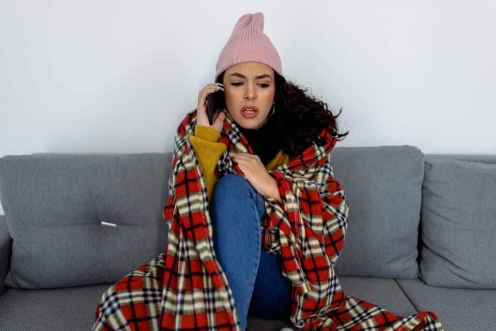 A woman in warm clothing feels cold and displeased while sitting at home, freezing, and calling Emergency HVAC Services on a mobile phone to get help.